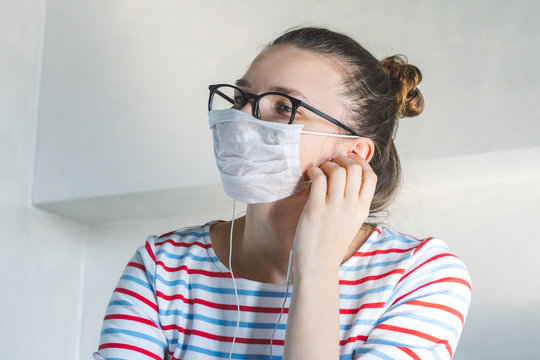 Close up of young woman in a surgical mask on face against SARS-CoV-2. quarantine and isolation at home because of coronavirus flu virus. sick female student listening to music in earphones.