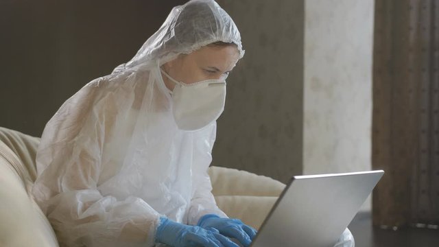 Woman in protective suit, protective mask and blue gloves works on a laptop during the quarantine.