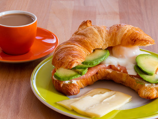 Croissant with salmon, poached egg and avocado on a wooden table with cup of coffee.