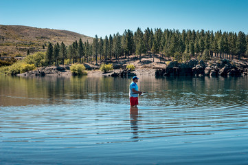 Fototapeta na wymiar Fisherman inside the lake with mountains and pine trees in the background. Neuquen, Argentina....