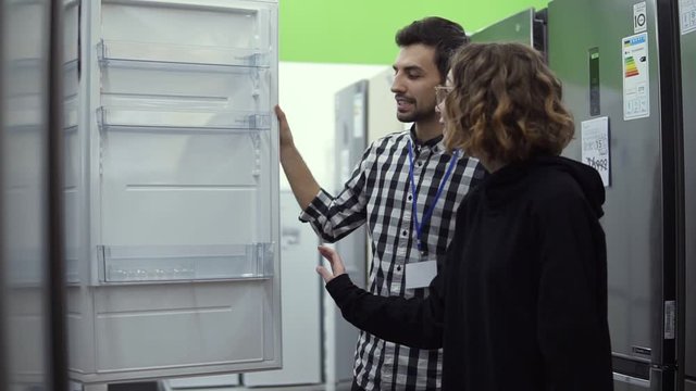 Young woman standing opening door of refrigerator with male consultant discussing design and quality before buying in a consumer electronics store. Discussing characteristics with store advisor