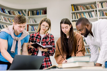 Beautiful young people in casual clothes, students, friends, working together in modern library, using a laptop, talking and smiling while making notes