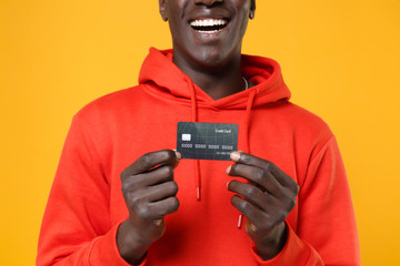 Cropped image of laughing young african american man guy in red streetwear hoodie posing isolated on yellow background in studio. People lifestyle concept. Mock up copy space. Hold credit bank card.