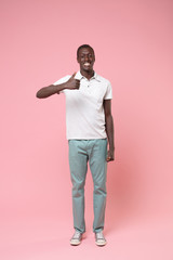 Smiling young african american man guy in white polo shirt, turquoise trousers posing isolated on pastel pink wall background in studio. People lifestyle concept. Mock up copy space. Showing thumb up.