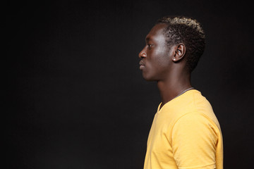 Side view of young african american man guy in yellow t-shirt posing isolated on black wall background studio portrait. People sincere emotions lifestyle concept. Mock up copy space. Looking aside.