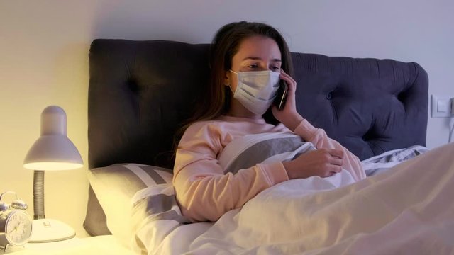 Sick woman with medical mask on the face lying in bed at home, coughing and calling a doctor.