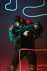 bi-racial cyberpunk man in protective mask aiming gun near neon lighting on black