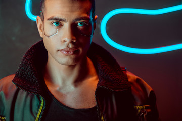 handsome and bi-racial cyberpunk man looking at camera near neon lighting on black