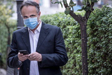 Manager in  giacca e camicia  indossa un mascherina azzurra e manda un messaggio dal suo telefono...