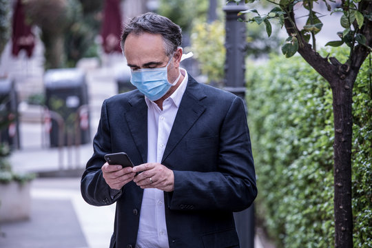 Manager in giacca indossa un mascherina chirurgica e utilizza il suo cellulare vicino a una siepe 
