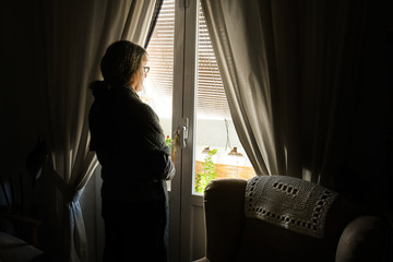 Mujer adulta, confinada por la pandemia del coronavirus COVID-19, mira hacia la calle a través de la ventana de su casa - obrazy, fototapety, plakaty