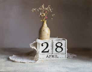 April 28, Vintage natural calendar. Day 28 of month, Handmade wood cube calendar with date month and day. Dry little roses in a vintage vase stand on the calendar on which lies a knitted napkin.