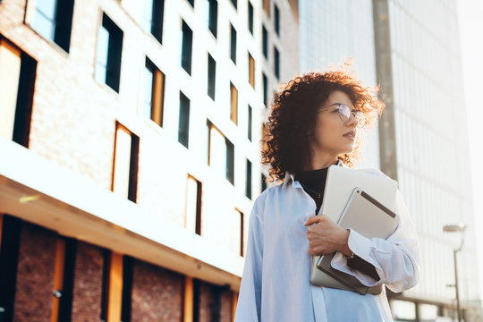 Lovely Business Woman With Curly Hair And Eyeglasses Is Walking Outside Carrying A Laptop And Tablet