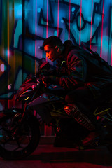 Obraz na płótnie Canvas side view of mixed race cyberpunk player riding motorcycle on street with graffiti