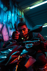 selective focus of mixed race cyberpunk player on motorcycle aiming gun on street with graffiti