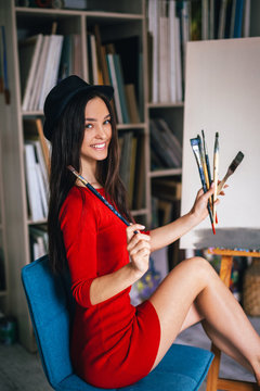 Woman Artist Sits On A Blue Chair Near An Easel With Brushes In Her Hands