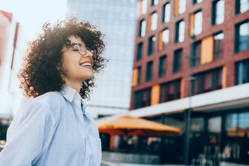 Cheerful woman with curly hair is cheering outside while wearing a white shirt and eyeglasses