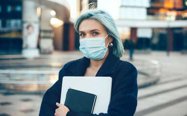 Blue haired caucasian entrepreneur holding a laptop while wearing a special white mask and looking...