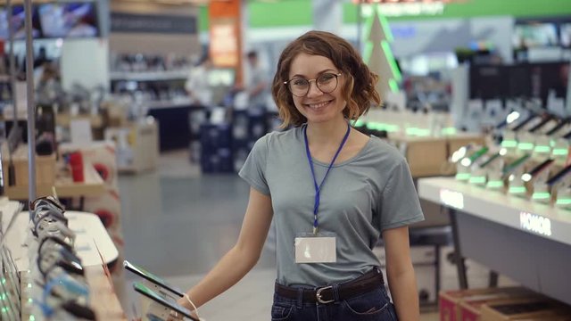 Positive female seller or shop assistant portrait in supermarket store. Woman in blue shirt and empty badge looking at the camera and smiling. Electronic devices on the background. Slow motion