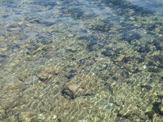Through the clear water in the rays of the reflection of the sun, you can see the shallow sea floor of sand, pebbles and stones of a gray brown color.