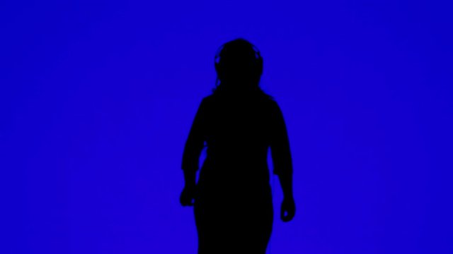 Silhouette of a slender woman wearing headphones and dancing to the music on a blue background