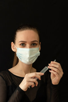 Beautiful girl in a protective mask and with a syringe in her hand. Protect yourself from the virus. Black clothes and background. Protection.