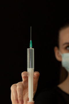 Beautiful girl in a protective mask and with a syringe in her hand. Protect yourself from the virus. Black clothes and background. Protection.