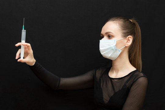 Beautiful girl in a protective mask and with a syringe in her hand. Protect yourself from the virus. Black clothes and background. Protection.