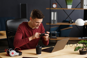 Young man playing games on mobile phone in home office with laptop. Relaxation, taking a break. Young people working with mobile devices. © YURII MASLAK