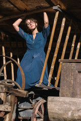 Fototapeta na wymiar A brunette woman wearing a cyan blue polka dot dress, black glasses and large hooped gold earrings is kneeling on an old wooden cart