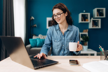 Cheerful girl with curly hair in casual clothing and eyewear working on laptop at living room and drinking coffee. Concept of modern technology and leisure time.