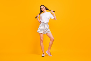 Full length photo of funny playful lady enjoy sunny day hold long hairdo hands making pigtails wear casual white t-shirt striped shorts shoes isolated vibrant yellow color background