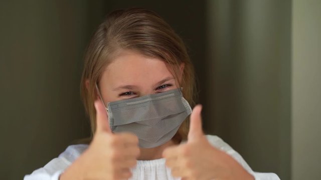 Close-up of a caucasian pretty child girl in protective medical mask looking at the camera, smiles and gives a thumbs up