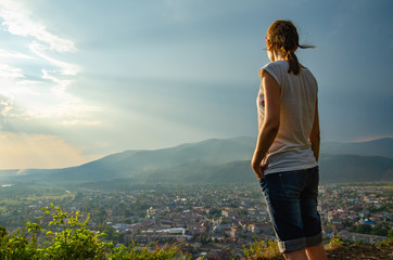 Young woman standing on a hilltop against the backdrop of an epic sky above a small town in a valley among the mountains