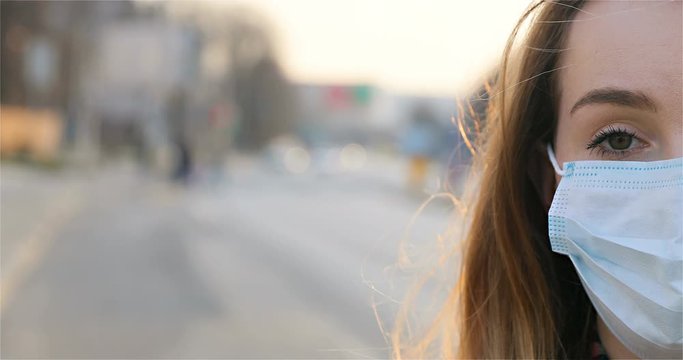 Close up of half face of sad Caucasian young woman in medical mask looking straight to camera while standing outdoor in city. Portrait of sick girl in protection and with upset look at empty street.