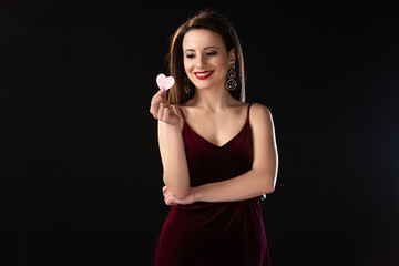 Fototapeta premium smiling woman in dress holding heart-shaped card isolated on black
