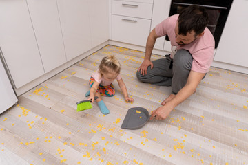 Cute little girl helps her father with cleaning the floor from cornflakes at the kitchen. Concept...