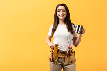 smiling handywoman holding metal cup and looking at camera isolated on yellow