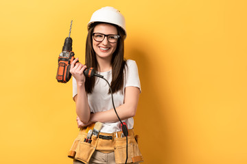 smiling handywoman in helmet holding drill on yellow background