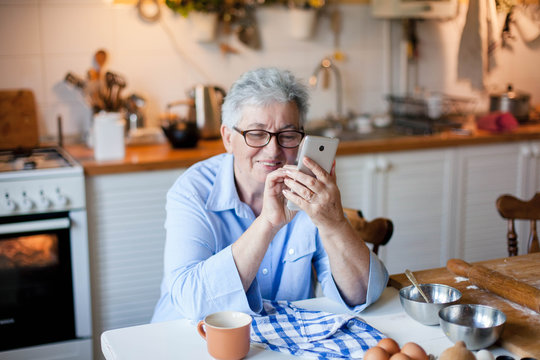 Senior woman using mobile phone at cozy home kitchen. Retired person shopping online. People connection, communication with gadget, delivery in isolation, distance healthcare. Lifestyle moment.