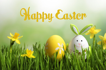 Colorful eggs with narcissus flowers in green grass and text Happy Easter against blurred background