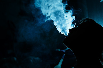 Silhouetted by a girl who smokes a hookah at night in a nightclub. It releases thick smoke from the...