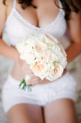 Obraz na płótnie Canvas Beautiful bride in a white lingerie holds a wedding bouquet closeup. Young caucasian woman sitting on a bed in lace underwear.
