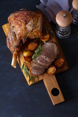 Roast leg of lamb with potatoes and rosemary on dark background. Overhead, flat lay, vertical image