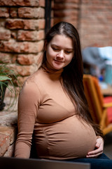 Portrait of a young pregnanty woman freelancer using laptop computer for distance job while sitting in modern coffee with cup of tea