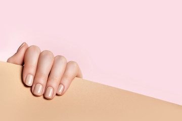 Female hand holds a sheet of paper and demonstrates a nude manicure. Pink, beige background with...