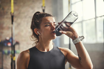 Fototapeta na wymiar Close up of young athletic woman in earphones is drinking water while exercising in gym.