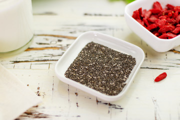 Close-up. Chia seeds lie in a minimalistic square saucer oon an old table . In the background are goji berries and fresh milk.