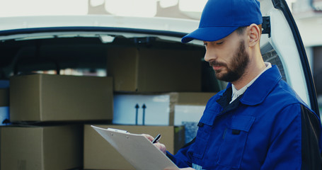 Close up of the Caucasian attractive male worker writing a document at the van full of carton parcels. Outside.
