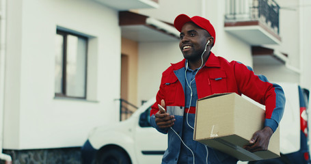 Fototapeta na wymiar Cheerful young African American deliveryman in red uniform taking out carton box from a van and walking to the house while listening to the music in headphones and smiling. Outdoor.
